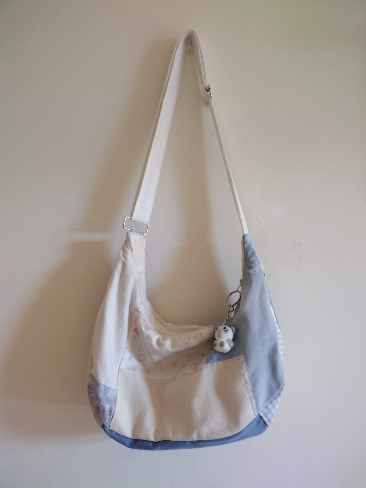 A small crescent-shaped bag made with patches of fabric, in beige, blue, and purple. A small teal panda keychain is hanging off of its zipper.