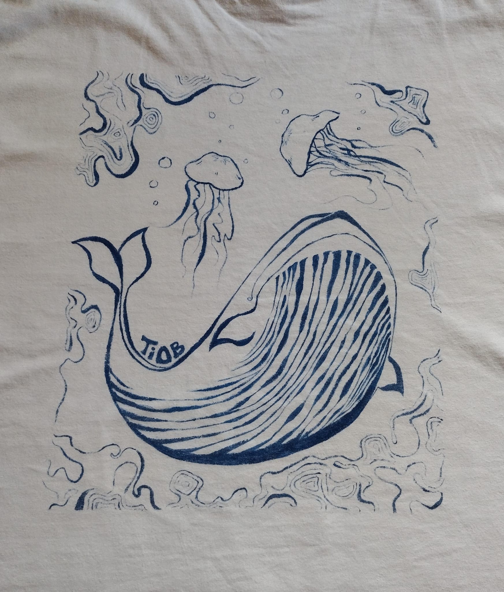 An image of the back of a white T-shirt, with a blue painted design I made. Design includes a large whale, two jellyfish, and a water motif.